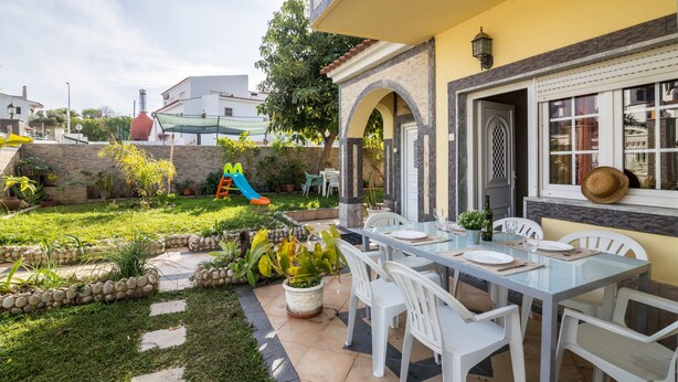 A patio with a table set for dining, surrounded by white chairs, and a play area with a slide in the background. - Apartment "Anastácio" with Garden View, Shared Pool, Wi-Fi & Balcony (Olhos de Água)