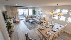 A spacious living room with a large sectional sofa, a dining table set for six, and a view of the outdoors through large windows. - Red Rock Views - 3 King En Suites + Rooftop Patio (St. George)
