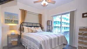 Une chambre à coucher comprenant un lit, une table de chevet, une lampe, une fenêtre donnant sur un immeuble et un ventilateur de plafond.