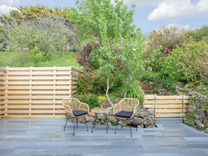 An outdoor patio with two wicker chairs and a small table, surrounded by a wooden fence and lush greenery.