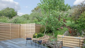 A wooden deck with two wicker chairs and a small table, surrounded by a wooden fence and lush greenery.