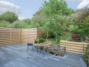 A wooden deck with two wicker chairs and a small table, surrounded by a wooden fence and lush greenery.