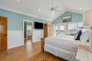 A bedroom with a large bed, wooden flooring, a ceiling fan, a television, and a view of the city through the window. - 8BR Beach Oasis w/ Pool & Rooftop (Isle of Palms)