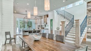 A spacious dining area with a wooden table and chairs, a kitchen in the background, and a staircase with a blue railing. - Luxury Home 1 Minute to the Gulf, with LSV, Bikes, and Heated Pool (Santa Rosa Beach)