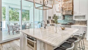 A modern kitchen with a large island, stainless steel appliances, and a view to an outdoor patio. - Luxury Home 1 Minute to the Gulf, with LSV, Bikes, and Heated Pool (Santa Rosa Beach)