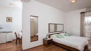 Une chambre d’hôtel avec un plancher en bois, un lit avec des oreillers verts, une table de chevet avec une lampe, un miroir, une fenêtre avec des rideaux et une kitchenette équipée d’un évier et d’une chaise.