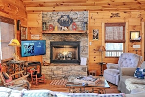 Un interior de cabaña acogedor con chimenea de piedra, paredes de madera, televisor y muebles rústicos.