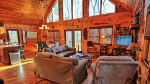 Ein gemütliches Blockhaushaus-Interieur mit Holzbalkendecke, einer Couch, einem Sessel, einem Esstisch und einem Fernseher.