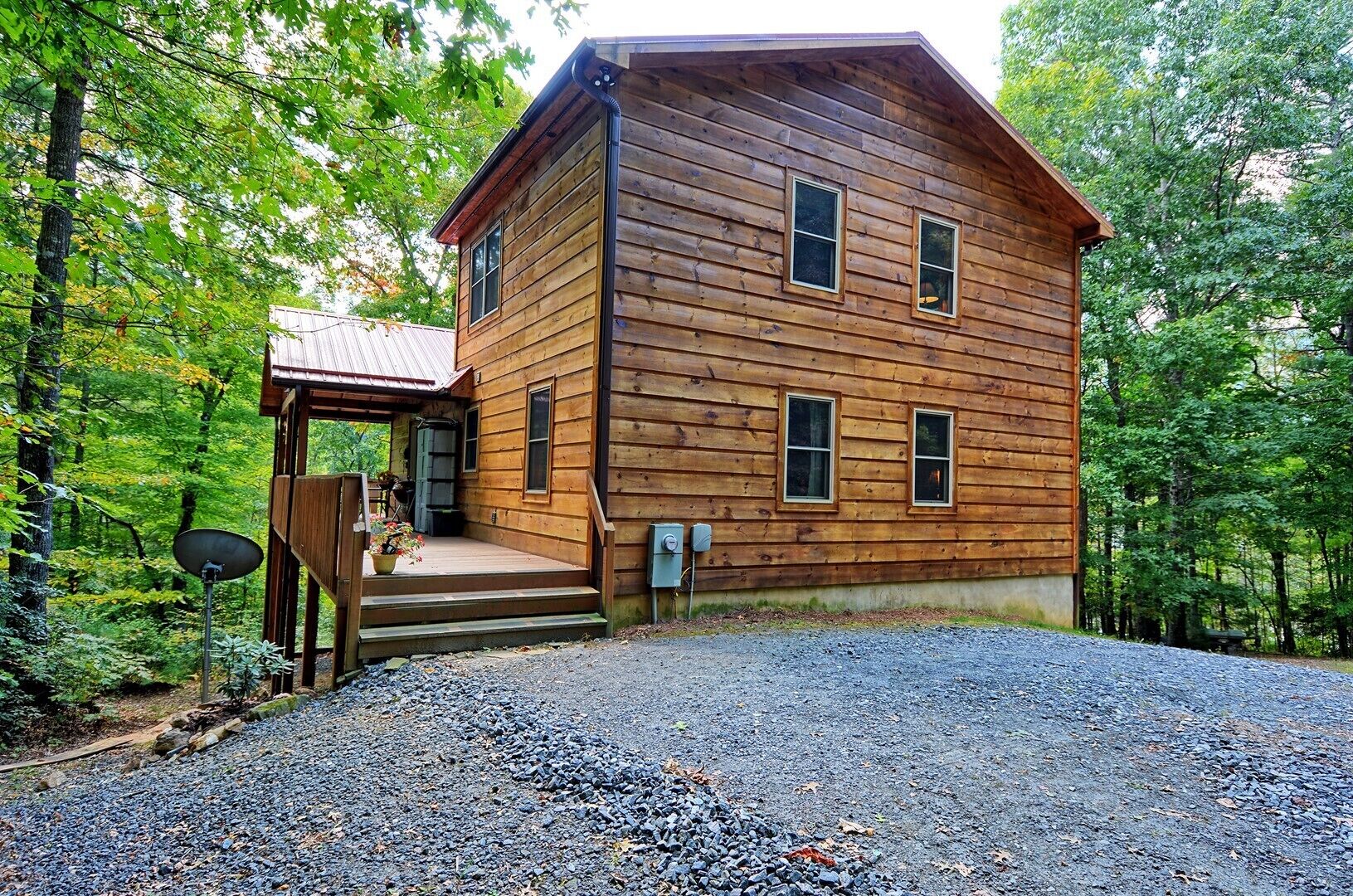 A wooden cabin with a porch, a gravel driveway, and a forested backdrop.