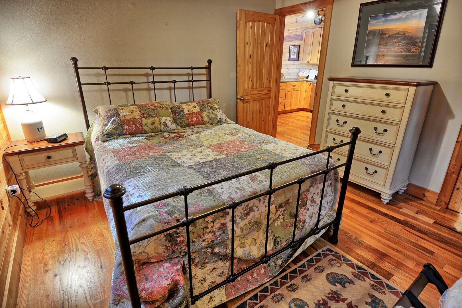 A bedroom with a metal bed frame, a wooden floor, a nightstand with a lamp, a dresser, and a rug.