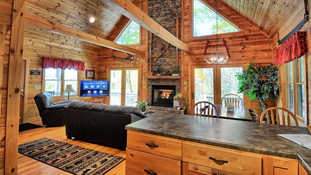 A log cabin interior with a fireplace, wooden ceiling beams, a stone wall, a dining area, and a television. - Spacious 3-bedroom cabin in West Jefferson, NC with game room and cozy fireplace. (West Jefferson)