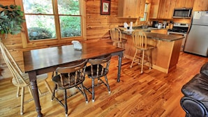 A wooden cabin interior with a dining table, chairs, a kitchen area, and a large window. - Spacious 3-bedroom cabin in West Jefferson, NC with game room and cozy fireplace. (West Jefferson)