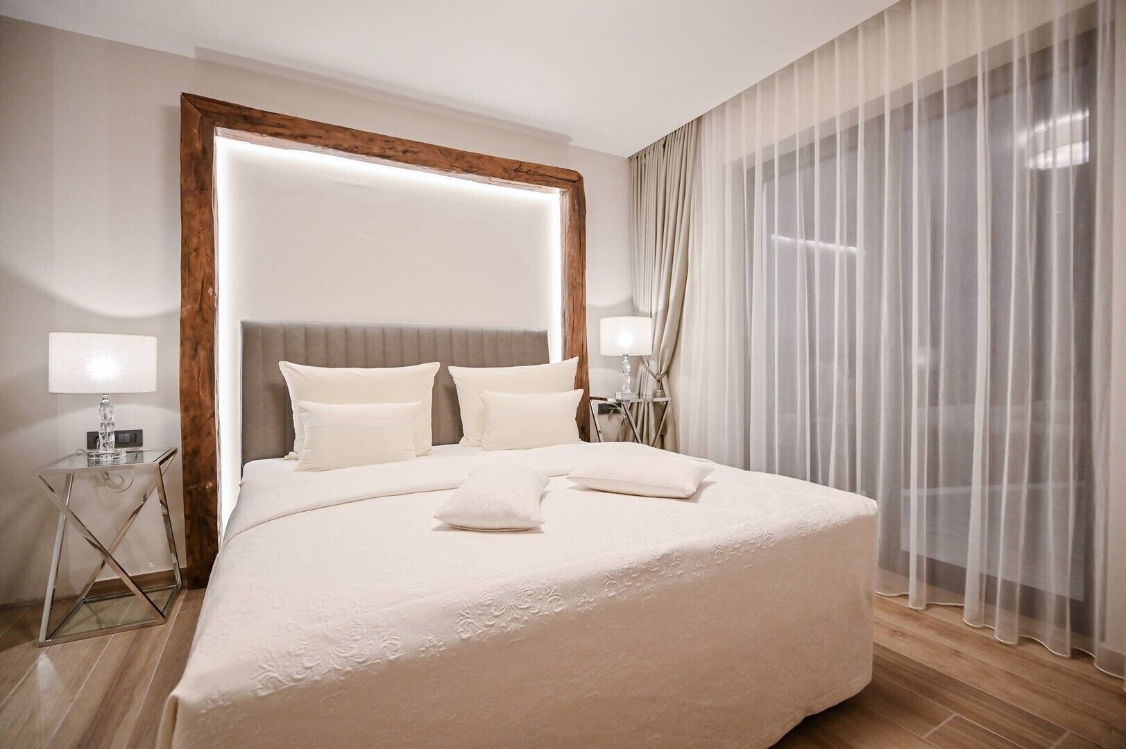 A modern hotel room with a large bed, two bedside tables, and a large window with sheer curtains.