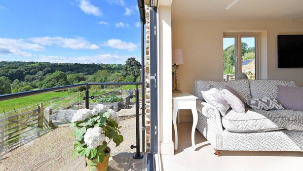Un balcón con vistas a colinas verdes, un sofá blanco con cojines estampados y una planta en maceta.