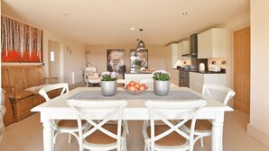 Un comedor con una mesa y sillas blancas, un cuadro en la pared y una cocina al fondo.