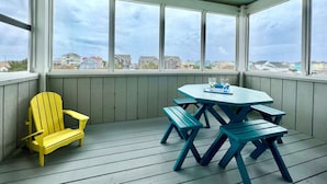 A covered outdoor area with a table and chairs, a yellow chair, and a view of houses through large windows. - N3325-Oceanside, Nags Head, Saturday Check In, Private Pool! (Nags Head)