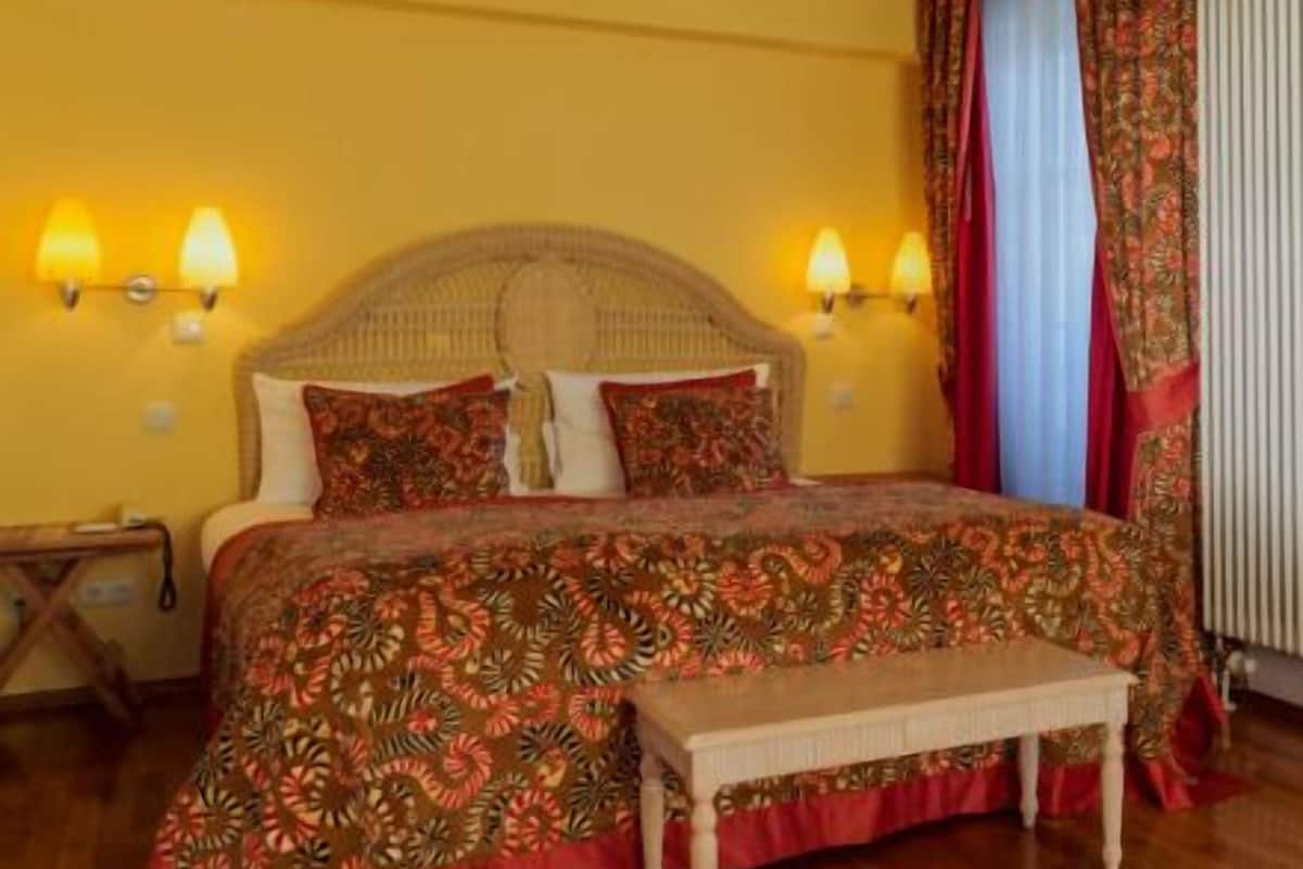 A bedroom with a bed, two bedside lamps, a nightstand, a table, and a window with curtains.