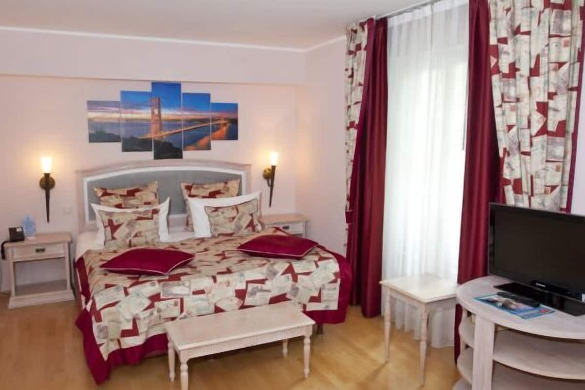 A hotel room with a bed, two bedside tables, a TV, and a window with curtains.