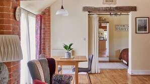 Een rustieke kamer met zichtbare houten balken, een bakstenen muur, een eettafel met stoelen en een gezellige fauteuil.