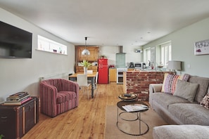 A modern living room with a sofa, a coffee table, and a striped armchair. A red refrigerator and a stove are visible in the kitchen area.