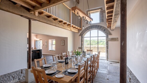 Uma sala de jantar com uma longa mesa de madeira posta para refeição, piso de pedra e vista através de uma janela em arco.
