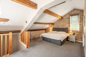 A bedroom with a sloped ceiling, exposed wooden beams, a stone wall, a bed, a chair, a nightstand, and a window with blinds. - Meadow Barn - Luxury Cottages (Sowerby Bridge)
