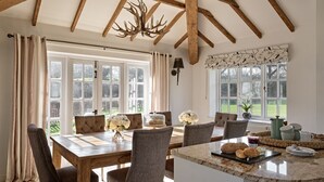 A dining area with a wooden table, upholstered chairs, a chandelier, and large windows with curtains. - Flock Cottage -  a cottage that sleeps 8 guests  in 4 bedrooms (Pudleston)
