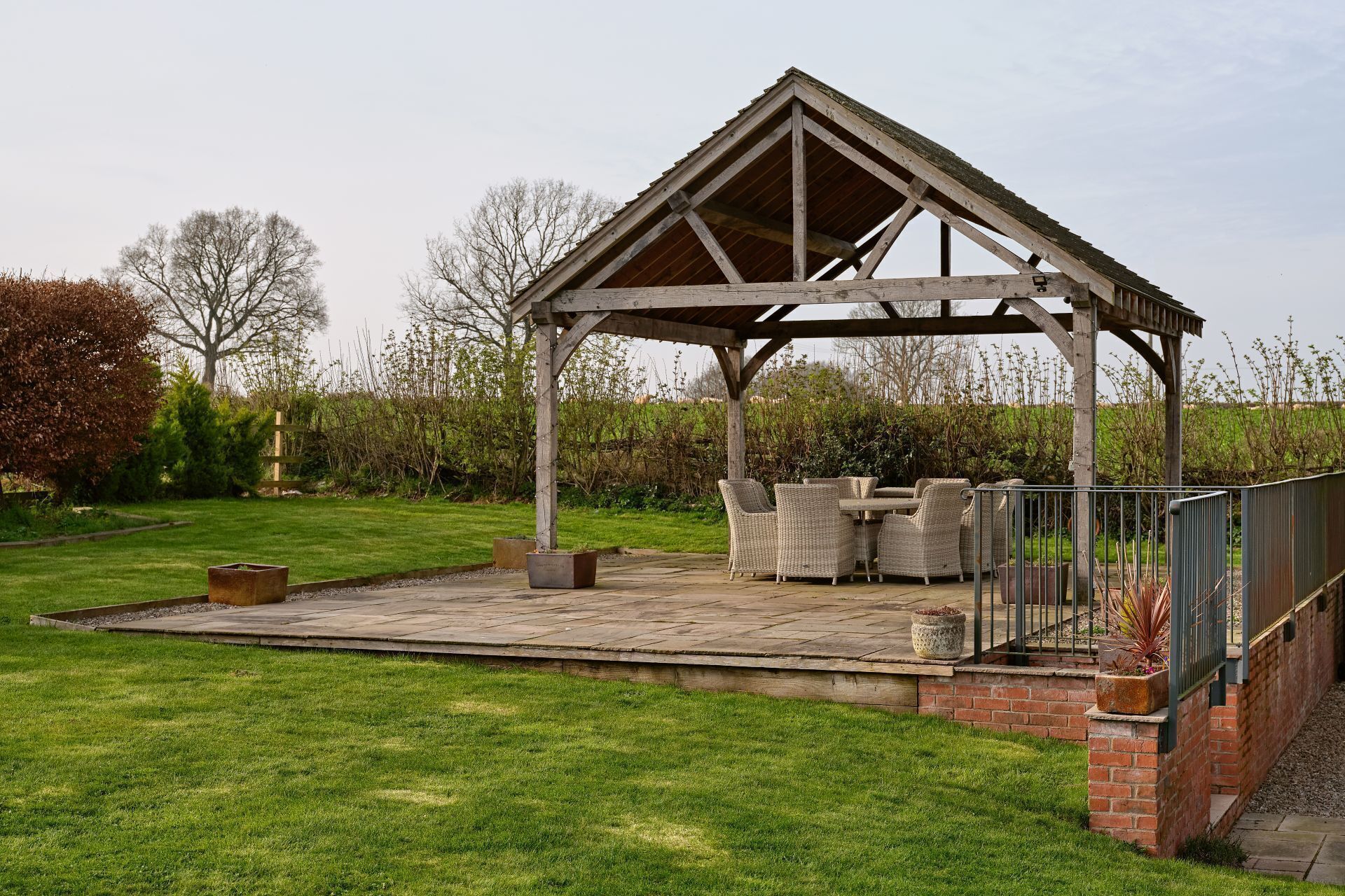 A covered outdoor seating area with a pergola structure, a brick patio, and surrounding greenery.
