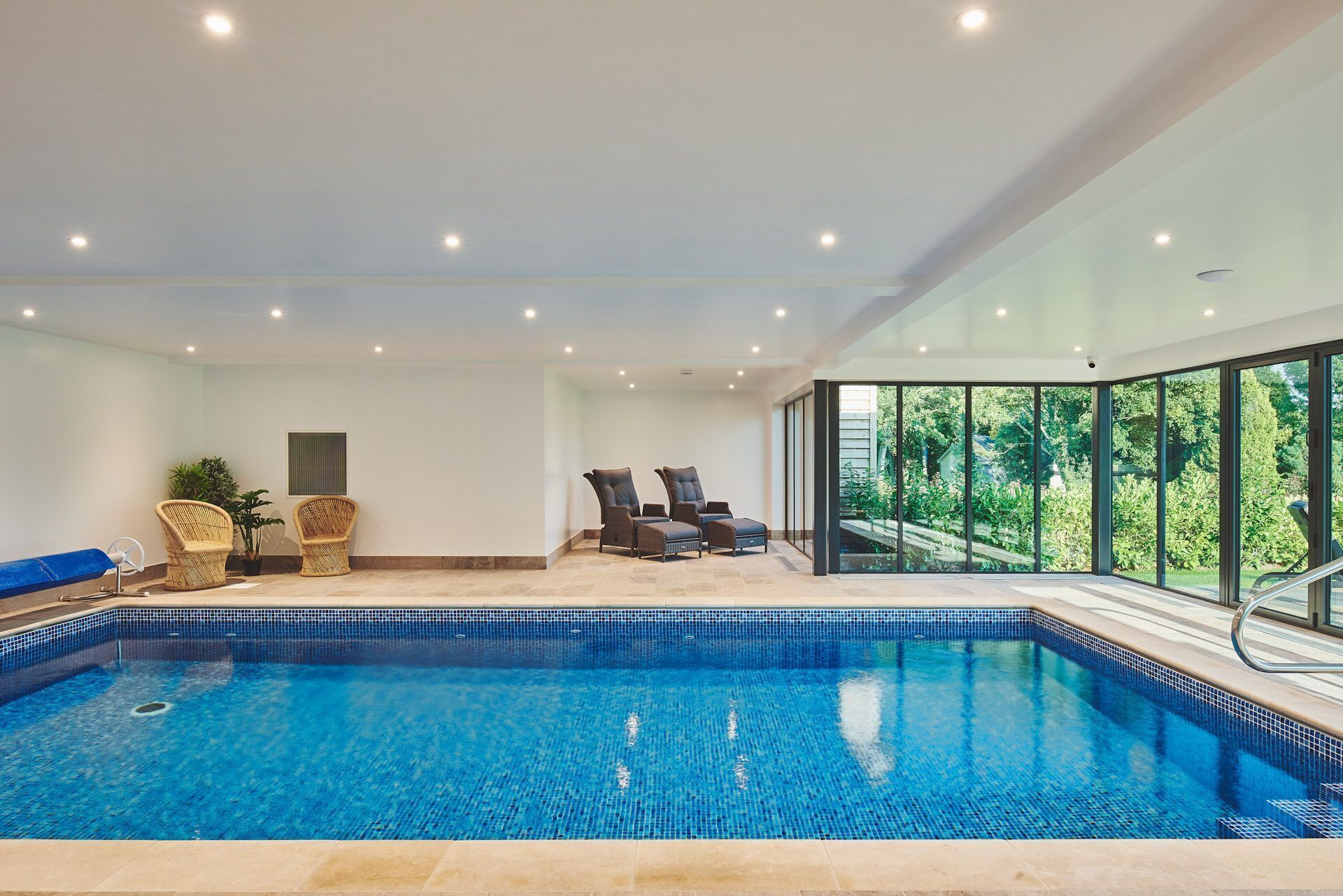 Ein moderner, überdachter Pool mit klarem, blauem Wasser, umgeben von Liegestühlen und einem durch eine Glaswand abgetrennten Bereich mit Blick auf Grünflächen.