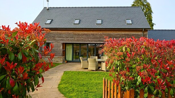 A house with a sloped roof, a patio with outdoor furniture, and a well-maintained lawn. - The Byre - Luxury Cottages (Leominster)