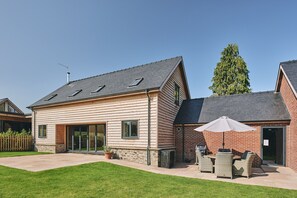 A two-story house with a sloped roof, large windows, and a patio with outdoor furniture. - The Byre - Luxury Cottages (Leominster)