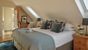 A bedroom with a sloped ceiling, a bed with multiple pillows, a bedside table with a lamp, and a nightstand with a book and a vase. - The Byre - Luxury Cottages (Leominster)