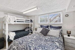 A bunk bed room with a patterned bedspread, a window with blinds, and a bedside lamp.