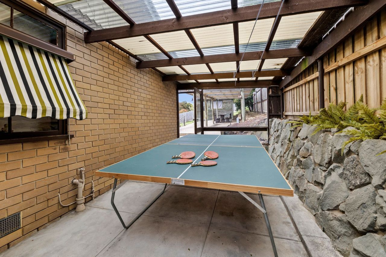 Une table de ping-pong installée à l’extérieur, avec deux raquettes, un mur en briques, un auvent en métal et un mur en pierre orné de verdure.