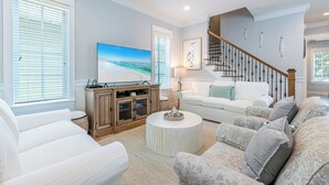 A living room with a large flat-screen TV, white sofas, a round coffee table, and a staircase with a wooden railing. - Screened Porch ~ Golf Cart ~ Community Pool (Miramar Beach)