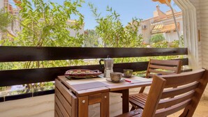 Una mesa de madera con una olla de metal y dos vasos, frente a una barandilla de balcón con vista a edificios y follaje.