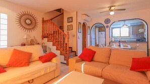 Una sala de estar con un sofá color beige, cojines rojos, una mesa de centro blanca, una escalera de madera y una cocina visible a través de un arco.