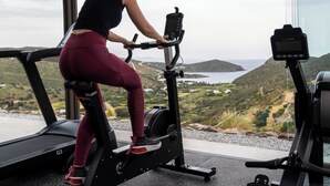 Uma mulher usando uma bicicleta ergométrica numa academia com vista panorâmica para colinas e um corpo d'água.