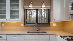 Une cuisine avec des armoires blanches, un comptoir en granit, un évier en acier inoxydable, une fenêtre donnant sur les arbres et une cuisinière à gaz.