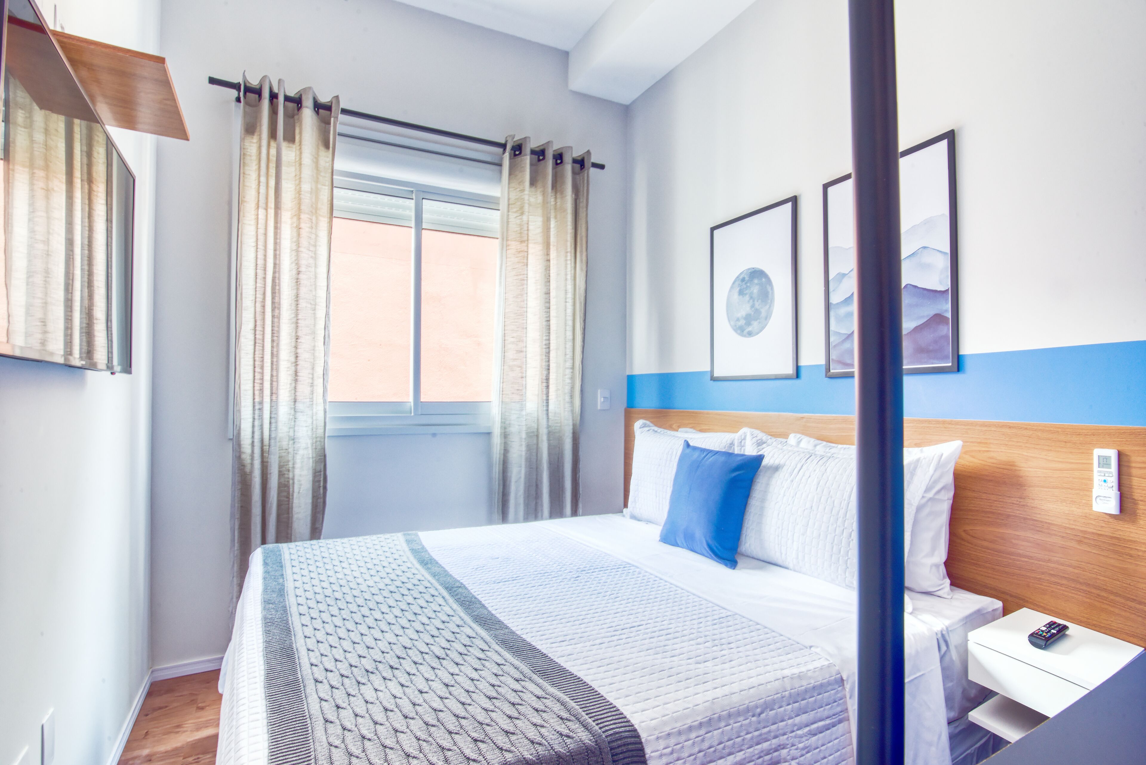 A hotel room with a large bed, a bedside table with a phone, and a window with curtains. There are three framed pictures on the wall.
