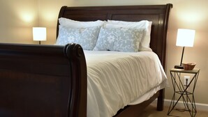 A bed with a dark wooden frame, white bedding, and two patterned pillows. There is a small table with a lamp and a basket beside the bed. - Spacious Townhouse in the ❤ of Fayetteville (Fayetteville)