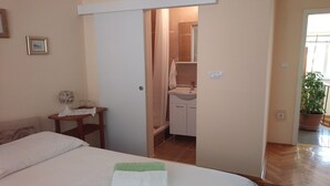 A bedroom with a bed, a bedside table with a lamp, a bathroom with a sink and mirror, and a potted plant in the corner. - Rooms Brezak - Double Room with Shared Kitchen (Lastovo)