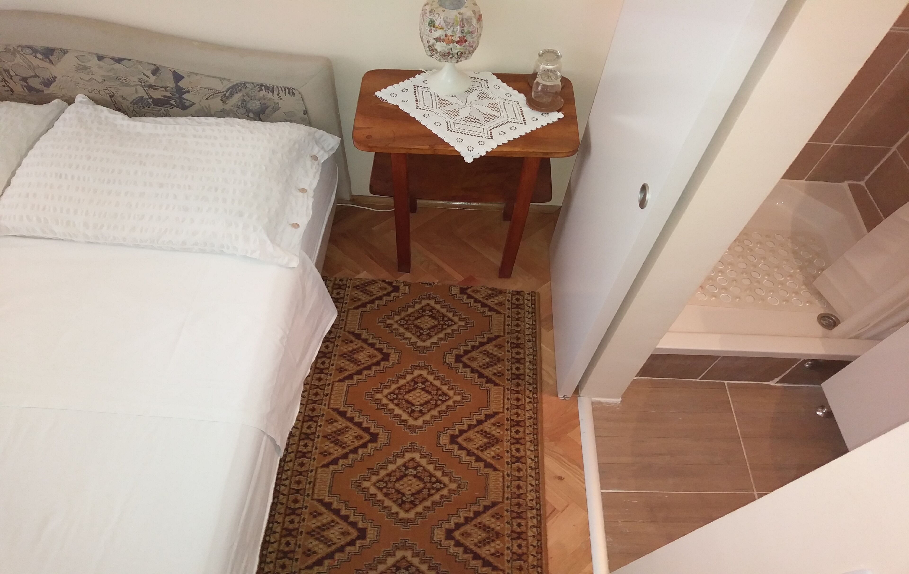 A bedroom with a bed, a small wooden table with a decorative item, a patterned rug, and a bathroom with a shower.