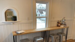 A bar area with a wooden counter, four metal stools, a small shelf with a sign, and a window with a view of a house. - Cozy Cottage located near Czech Village and Newbo areas in Cedar Rapids, IA (Cedar Rapids)