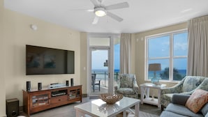 A living room with a flat-screen TV, a ceiling fan, a sofa, a coffee table, and a view of the ocean through large windows.