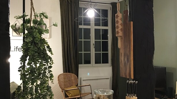 Un salon chaleureux avec une plante suspendue, un fauteuil en osier, une table en verre sur laquelle se trouvent un bol et un bocal rempli d’en-cas, et un téléviseur en arrière-plan.