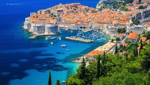 A coastal city with historic architecture, a harbor with boats, and a clear blue sea.