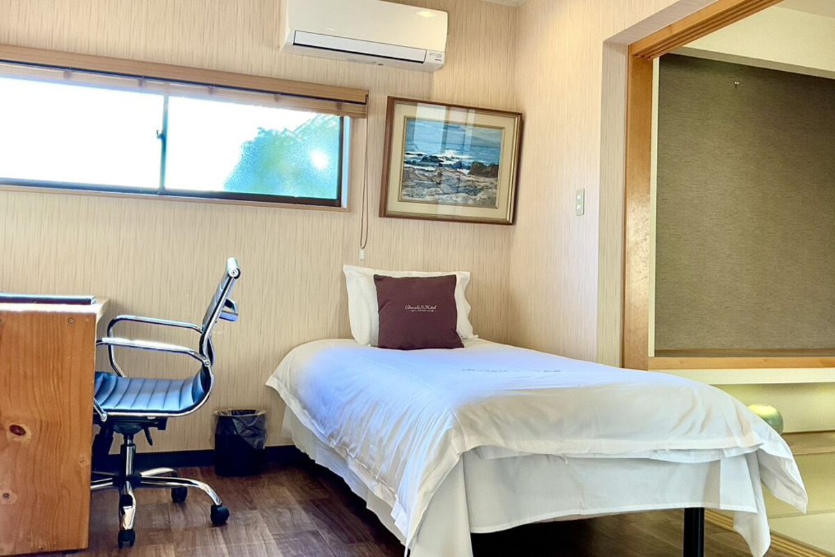 Habitació d'hotel amb un llit, una taula de treball, una cadira i una finestra gran.