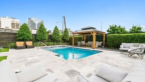 A swimming pool with a pergola, outdoor furniture, and a cityscape in the background.