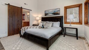 Un dormitorio con una cama grande, una mesita de noche, una lámpara, una ventana con persianas, un cuadro en la pared y una puerta de madera.